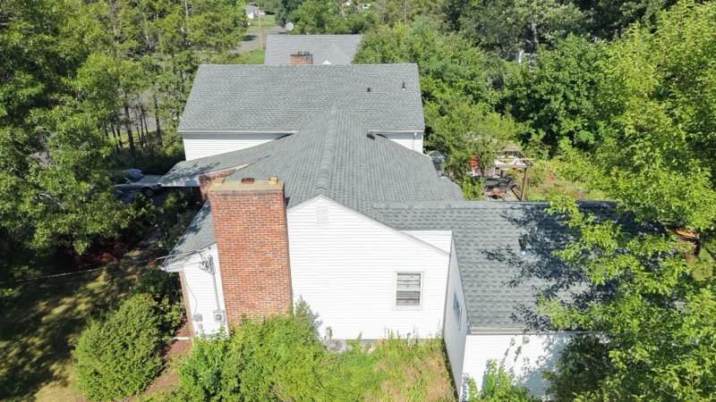 An aerial view of a white home’s shingled roof, with a brick chimney and trees behind it